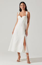 A woman is wearing a white sleeveless dress with wide shoulder straps, a sweetheart neckline, and fitted bodice. The dress has a slit on the right side, reaching up to mid-calf. She is also wearing strappy, open-toe high-heeled sandals.